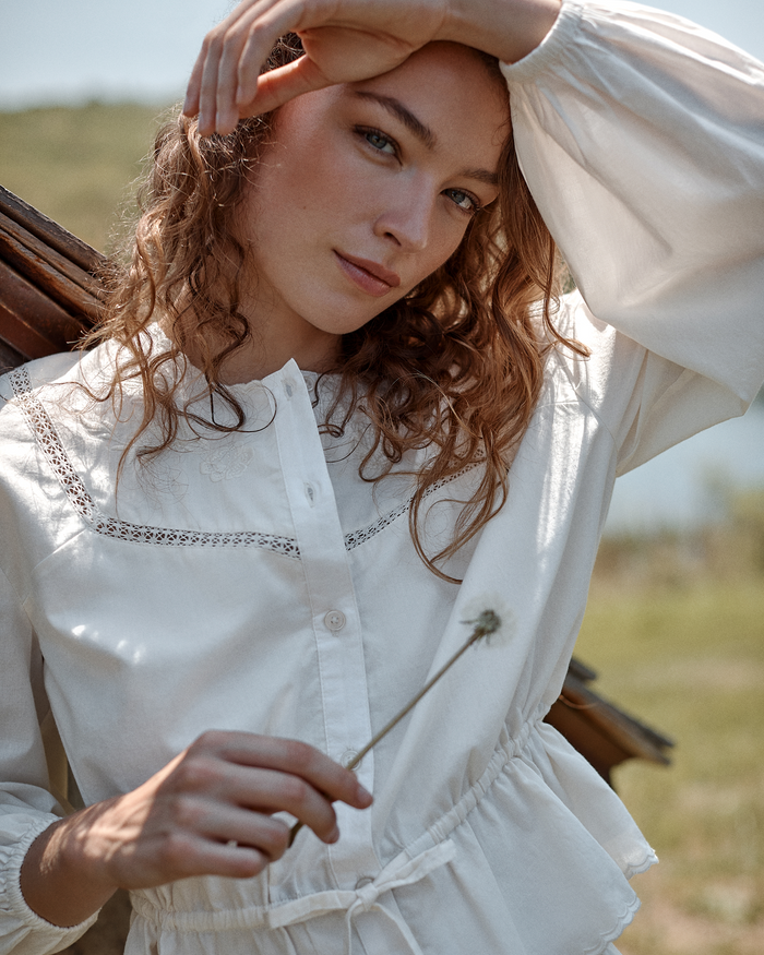 A woman wearing the Women’s Voile Marguerite Pajama Set in Ivory stands outdoors, holding a dandelion and shielding her eyes from the sun with her hand. She has curly hair and a serene expression, framed by a blurred natural background.
