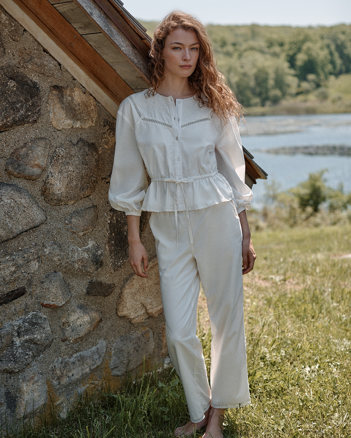 A woman wearing the Women’s Voile Marguerite Pajama Set in Ivory stands barefoot by a stone wall outdoors, with water and green trees in the background on a sunny day.