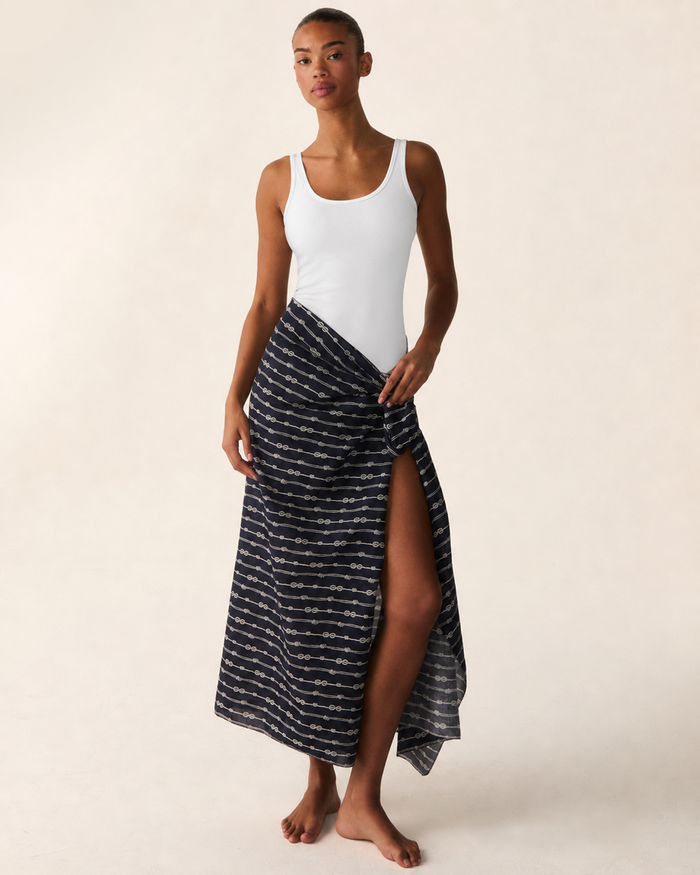 A woman stands barefoot against a light beige background, wearing a white tank top paired with the Women’s Voile Pareo in Nautical Knots—a navy blue patterned wrap skirt featuring a high slit.