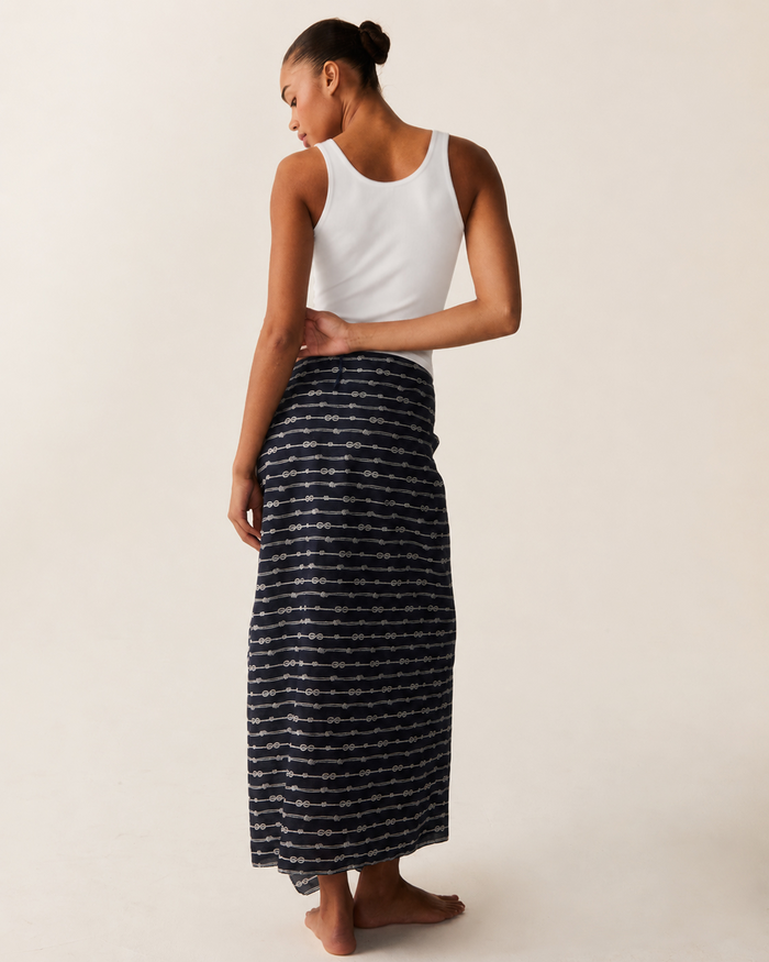 A woman stands barefoot, facing away, wearing a white sleeveless top and the Women’s Voile Pareo in Nautical Knots—a long skirt featuring a dark design with horizontal dotted patterns against a light background.